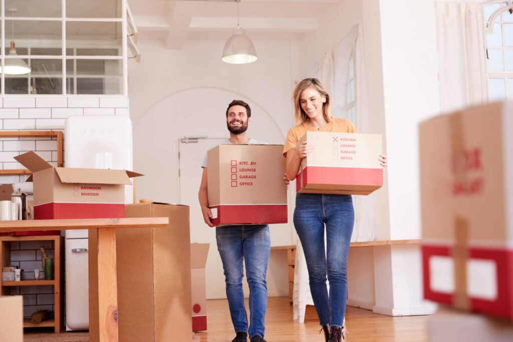 A man and a woman smile as they carry moving boxes in a bright room with large windows. More packed boxes are scattered around the wooden floor. The room has white brick walls and a modern interior design.