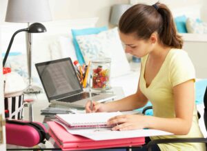 A young woman with a ponytail, wearing a yellow shirt, is sitting at a desk writing in a notebook. A laptop with an open document, various stationary items, and a jar of colorful candies are placed on the desk. The room has a cozy and organized setup.