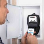 A man stands in front of an open electrical meter box, taking a photo of the meter's display with his smartphone. The meter shows the reading in black numbers on a white background. He appears to be documenting the reading.