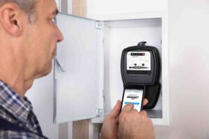 A man stands in front of an open electrical meter box, taking a photo of the meter's display with his smartphone. The meter shows the reading in black numbers on a white background. He appears to be documenting the reading.
