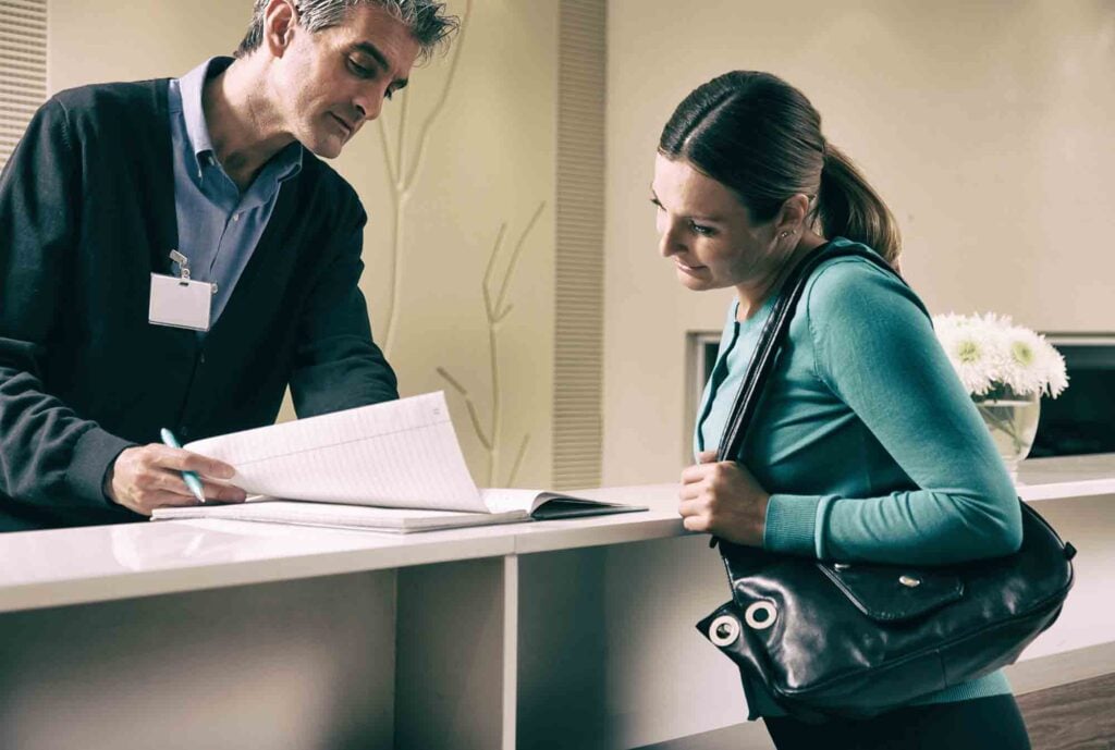 A man wearing a badge is assisting a woman at a reception desk. The woman, with a ponytail, is leaning on the counter, holding a black handbag and looking at the book the man is writing in. The background features minimalistic decor and a vase with white flowers.
