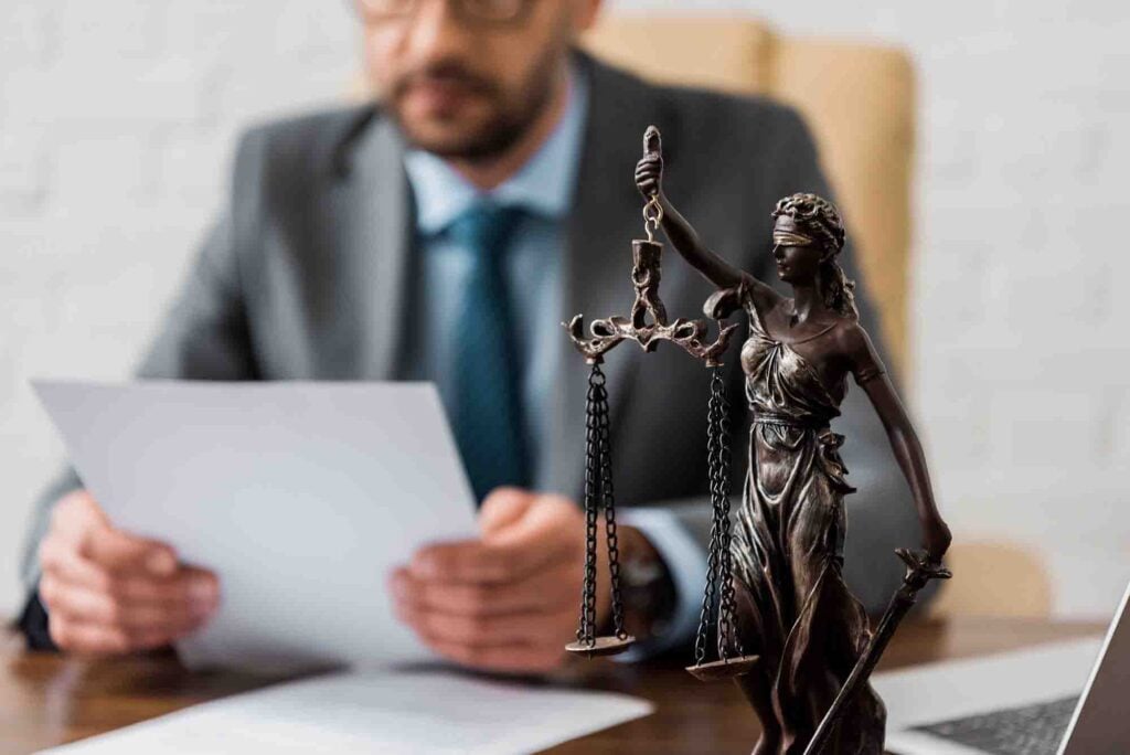A suited man sits at a desk reading a document, with a bronze statuette of Lady Justice in the foreground. The statuette holds scales and a sword, symbolizing fairness and law. The environment appears to be an office setting.