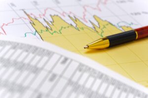 A close-up of a ballpoint pen resting on a sheet of financial data, including a line graph with red and green lines and a numeric table of values. The pen is gold and black with a red accent, placed diagonally across the chart.
