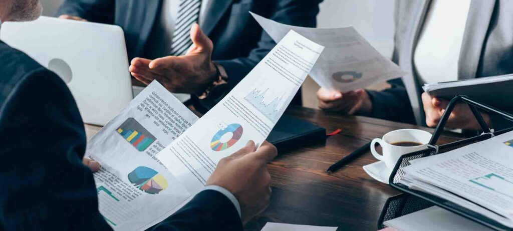 A group of professionals in business attire sitting around a table, holding and discussing documents with various charts and graphs. A laptop, cup of coffee, and more papers are on the table. Only the hands and partial torsos of the individuals are visible.