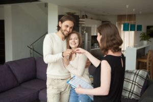 A smiling couple stands in a modern living room as a woman hands them a set of keys. The couple is dressed casually and showing excitement. The woman is holding a clipboard, suggesting a house or apartment agreement signing. The setting is bright and welcoming.
