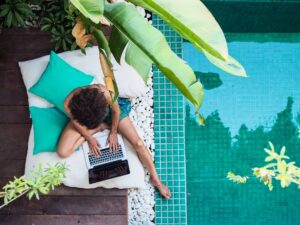 A person sits on a white cushion next to a turquoise swimming pool, working on a laptop. They are partially shaded by large green leaves. Two teal pillows are placed by their side, and white pebbles surround the wooden deck area.