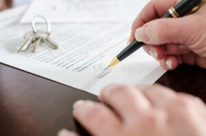 A person signing a document with a black and gold pen on a wooden surface. Nearby, a set of keys lies on top of the document, partially visible, suggesting the signing might relate to property or real estate.