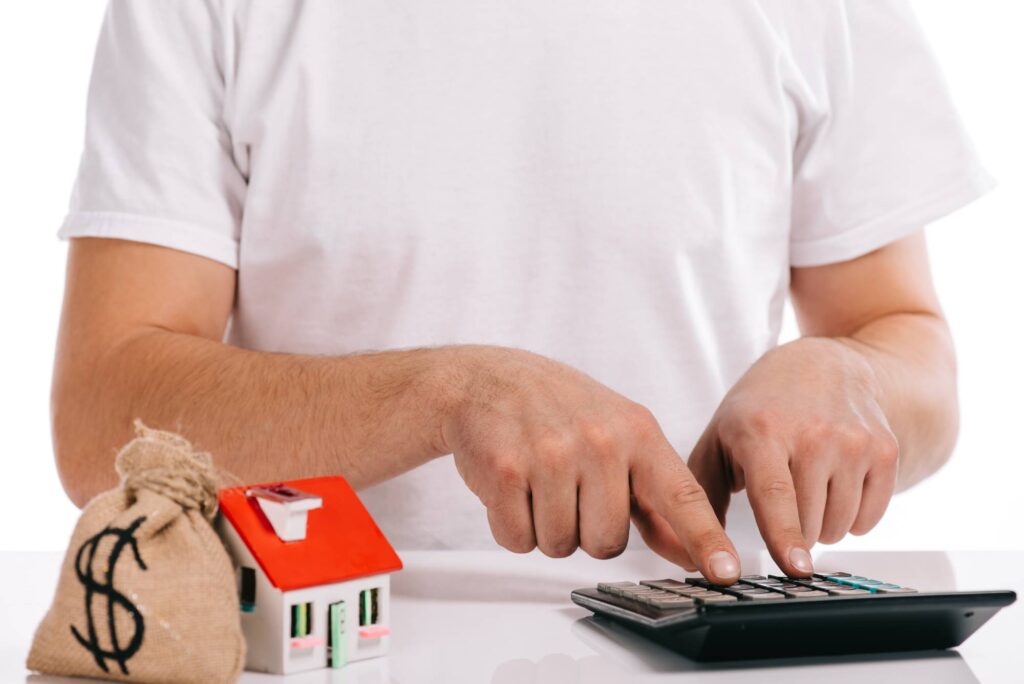 Person using a calculator with a small model house and a bag with a dollar sign on it nearby. The person is wearing a white shirt and the background is plain white.
