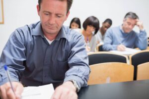 A group of adults sit in a classroom, writing on notepads. The man in the foreground is focused on his work, while a diverse group of individuals behind him are also engaged in writing. The classroom has wooden desks and a neutral-colored wall.