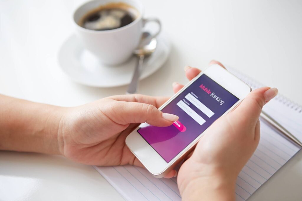 A person holds a smartphone with a mobile banking login screen displayed. They are using their finger to interact with the phone. A cup of coffee and a notebook with a pen are visible in the background on a white surface.
