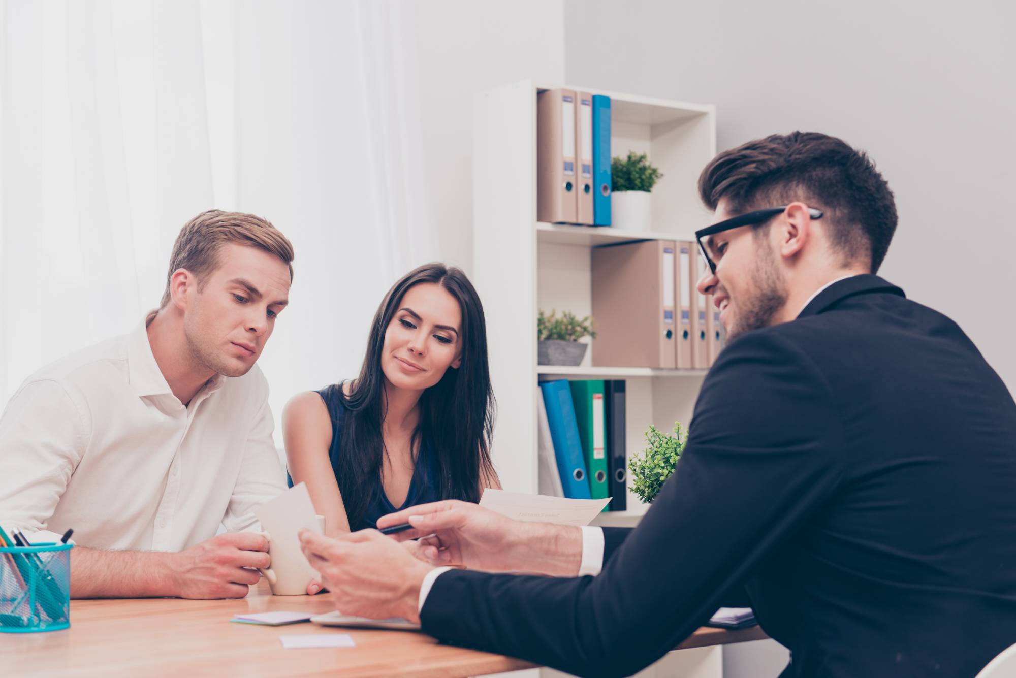 A man and a woman sit at a desk, attentively listening to another man in glasses who is explaining something to them. The setting appears to be an office, with shelves of folders and files in the background. The table has some documents and a small plant.