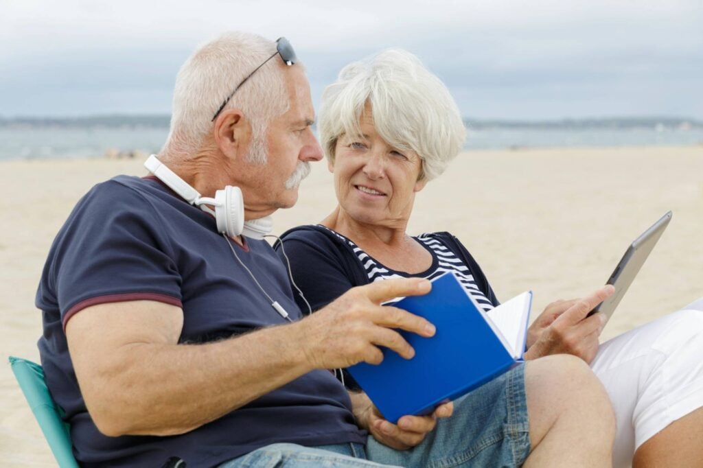 Expat couple sitting on the beach. The man is reading a book and the woman is reading an iPad