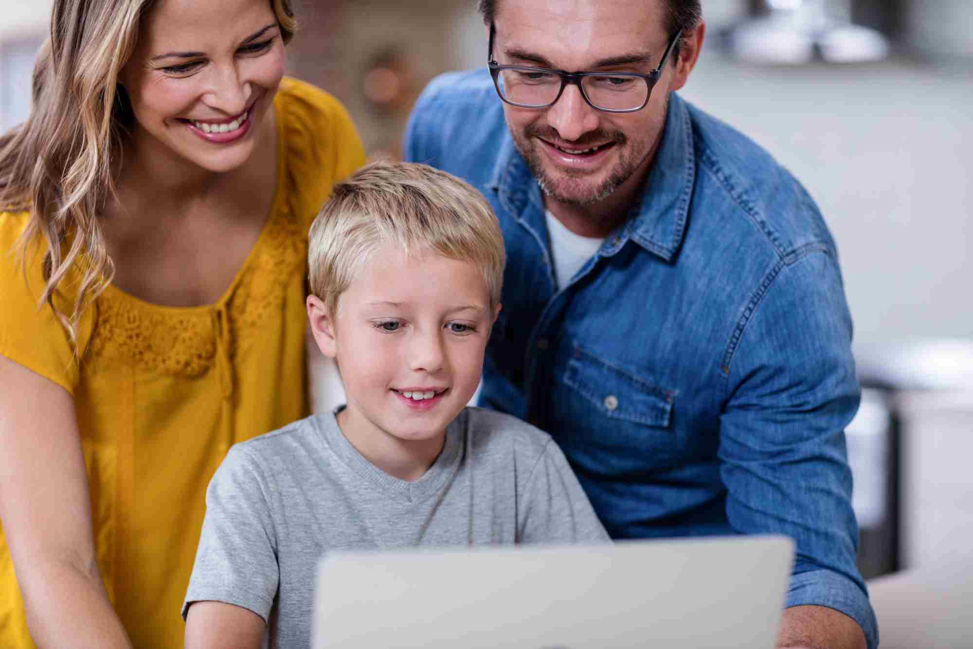 A smiling couple and their young son are looking at a laptop together. The woman is wearing a yellow top, and the man is in a blue denim shirt. The boy, who is seated, is focused on the screen. They appear to be enjoying their time together.