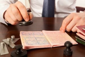 A person stamps a passport at a wooden desk. Several stamps and ink pads are scattered on the desk, along with a few documents. The person is wearing a white shirt and a blue tie.