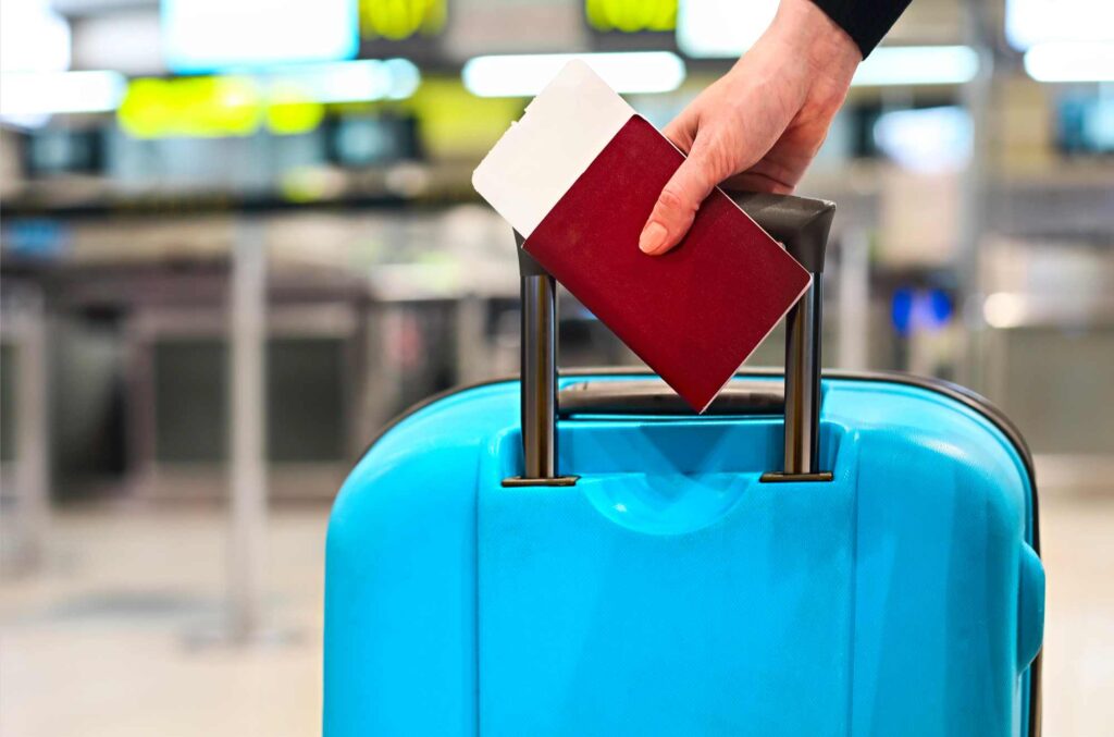 A person’s hand holding a passport and boarding pass rests on the handle of a blue suitcase at an airport check-in area, with blurred signs and counters in the background.