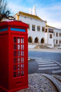 A bright red British-style telephone box stands on a cobblestone street corner, with a large historic white building featuring arched windows and unique chimneys in the background. The sky above is clear with a hint of clouds.
