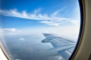View from an airplane window showing a blue sky with a few wispy clouds above and farmland below. The airplane's wing extends from the bottom right corner of the image. The window frame is partially visible.