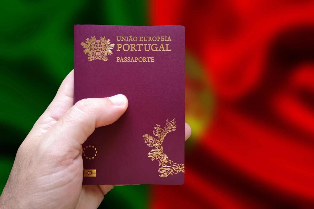 A hand holding a Portuguese passport in front of a blurred background resembling the colors of the Portuguese flag, green and red.