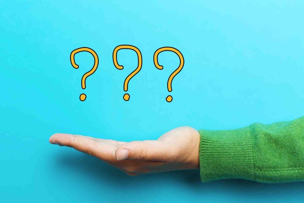 A hand in a green sweater is extended against a light blue background. Floating above the palm are three large, yellow question marks.