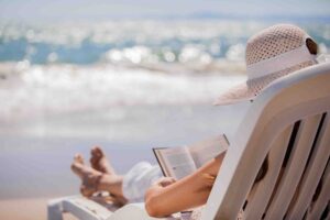 A person lounging on a beach chair, wearing a large sun hat and white clothing, is reading a book. In the background, gentle waves are visible under a bright blue sky. The sun is shining, creating a serene and relaxed atmosphere.