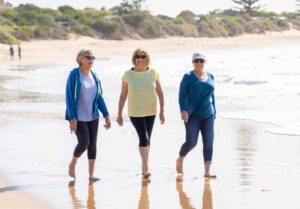Three elderly women walk along a beach shoreline on a sunny day. They are casually dressed in light jackets and pants, and they seem to be enjoying their time together. The ocean waves gently touch their bare feet, and the background features greenery and distant trees.
