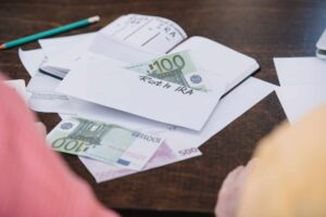 An envelope labeled "Roth IRA" with a 100 euro note partially visible, surrounded by various euro bills, documents, and a green pencil on a wooden table. Two people are partially visible on either side of the table.