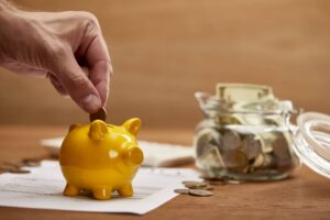 A hand puts a coin into a yellow piggy bank on a table, with money and papers, suggesting saving for life or moving to Portugal.