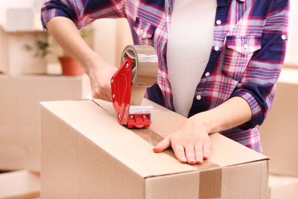 A person wearing a plaid shirt is sealing a cardboard box with packing tape using a tape dispenser. The scene appears to be indoors, with other boxes and a small potted plant blurred in the background.
