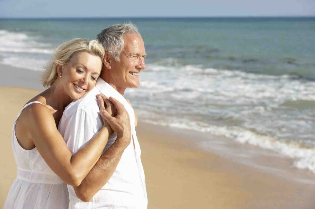 An elderly couple stands on a sandy beach by the ocean. The woman, in a white sleeveless dress, smiles while hugging the man, also in a white shirt, from behind. Both are joyful, looking out towards the water under a clear sky.