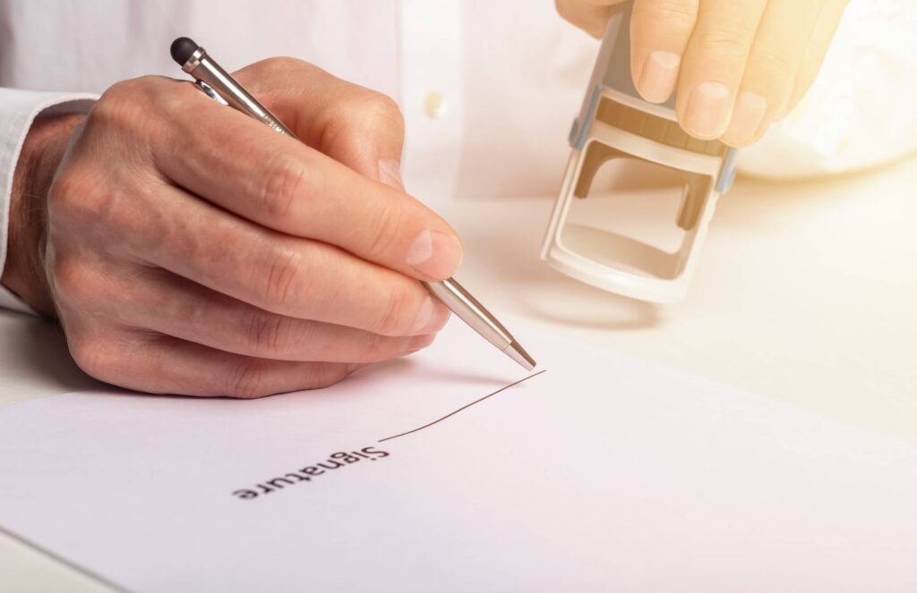 A person holding a pen is signing a document labeled "Signature" while another hand prepares to stamp the paper with a rubber stamp. The setting appears to be a well-lit desk or workspace.