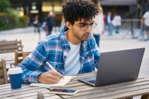 A person with curly hair and glasses is seated outdoors at a table, working on a laptop. They are writing in a notebook, with a smartphone and a coffee cup on the table. The background shows a blurry urban environment with people and buildings.