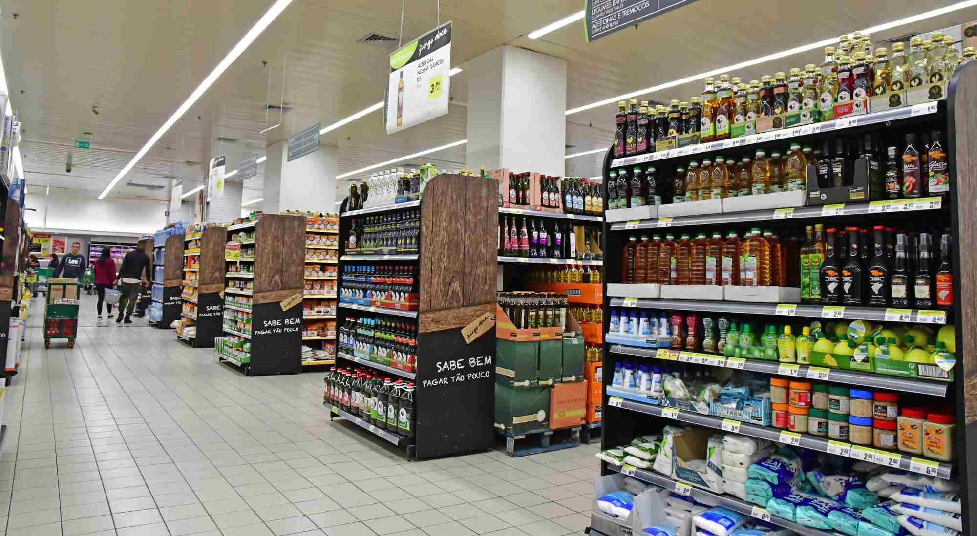 A supermarket aisle with well-stocked shelves displaying various products, including bottles of condiments, oils, and sauces on the right, and packaged food items on the left. Shoppers can be seen in the background with shopping carts. The floor is tiled.