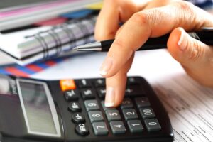 A close-up of a person's hand using a calculator. The person is holding a pen in one hand and pressing the buttons on the calculator with the other. In the background, there are documents, papers, and a notebook with a spiral binding.