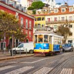A charming street scene in Lisbon, Portugal, with a vintage yellow tram labeled "28" traveling along cobblestone tracks. The backdrop features colorful buildings, including a red one, trees with autumn foliage, parked vehicles, and a quaint urban atmosphere.
