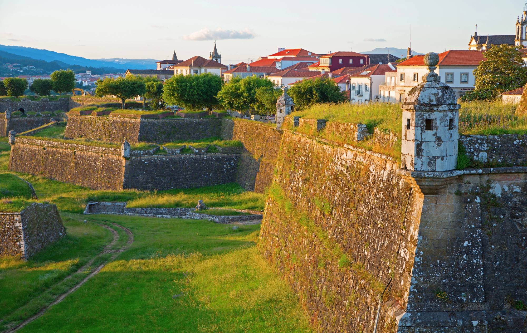 The image shows the historic fortifications of Valença, a European town in Portugal at sunset, featuring stone walls and manicured green lawns. The town in the background has a mix of white and red-roofed buildings, with distant hills, adding to the picturesque scenery.