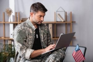 A man in a camouflage uniform and dog tags sits indoors using a laptop, with an American flag nearby; shelves are in the background.