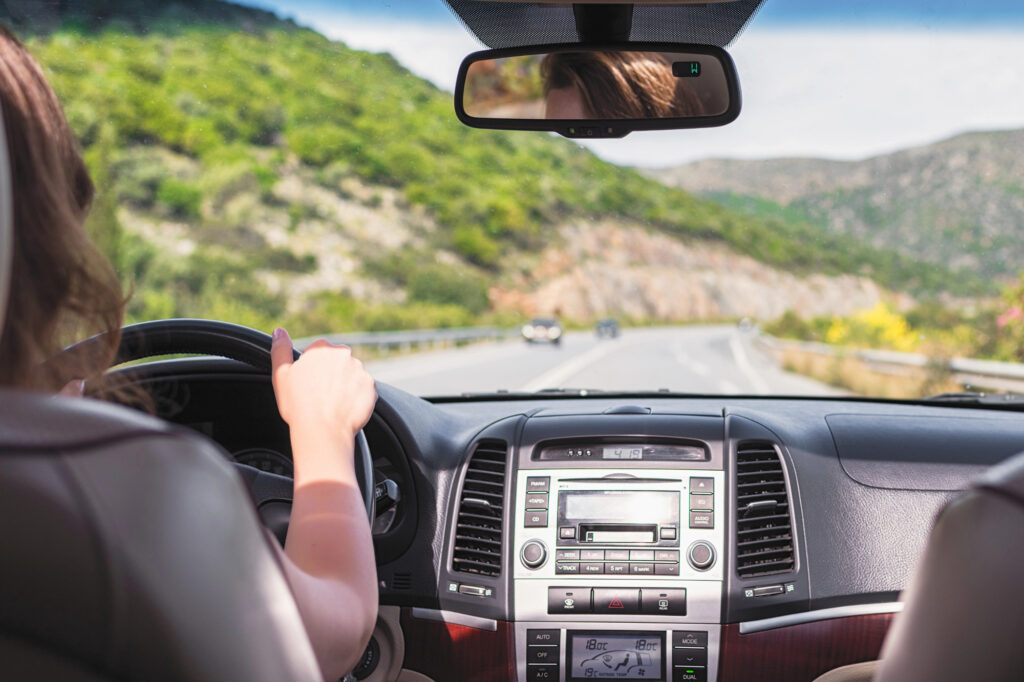 A woman is driving in a car on a two-lane road with cliffs on the left-hand side