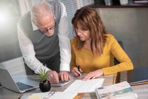 Two people are working together at a desk. An older man with gray hair and glasses is standing and operating a calculator while a woman with long hair in a yellow sweater is seated, holding a pen and looking at documents. A laptop and plant are on the desk.
