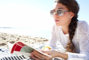 A woman with sunglasses and a braid is lying on a striped blanket at the beach, reading a magazine. She is wearing a white sweater and appears to be relaxed, with the sea and sky in the background.