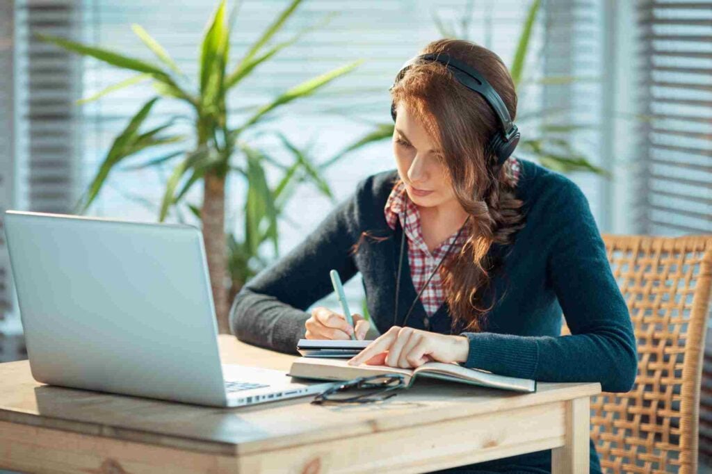 A woman wearing headphones sits at a wooden table in front of a laptop. She is writing in a notebook with a pen. The background features large green plants and window blinds that let in natural light. Glasses and other items are placed on the table.