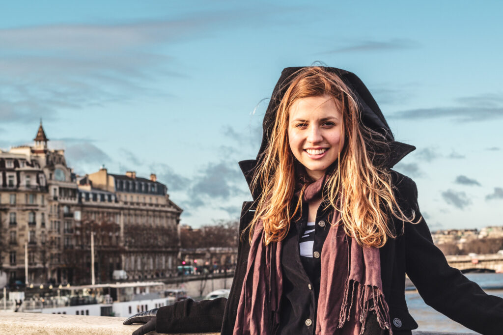 A young woman in a black coat and scarf smiles on a bridge, with historic buildings and a river in the background under blue sky.
