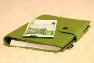 A green felt book cover with a button closure lies on a soft beige surface. A folded 100 euro banknote is placed on top of the cover. A small black tag with white text is visible on the book cover's side.