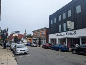 A street view of a small town with various shops and parked cars. On the right, there is a black building housing "Alexandra's Boutique". Further down the street are more businesses and a church steeple visible in the distance. The sky is overcast.