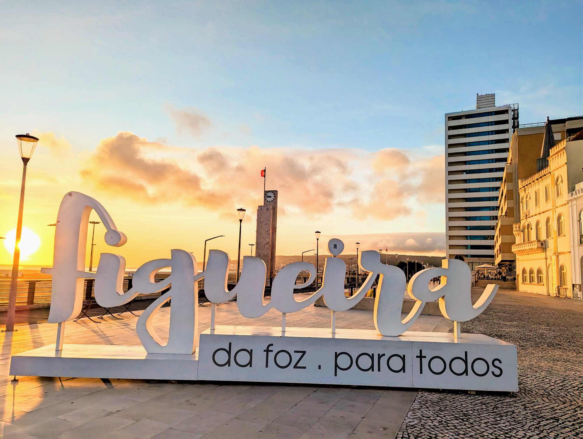 A large, white sign reads "Figueira da Foz para todos" with a backdrop of a modern, tall building and an older, ornate structure in Portugal. The sky is clear with a few clouds, and the setting sun casts a warm, golden light over the scene.