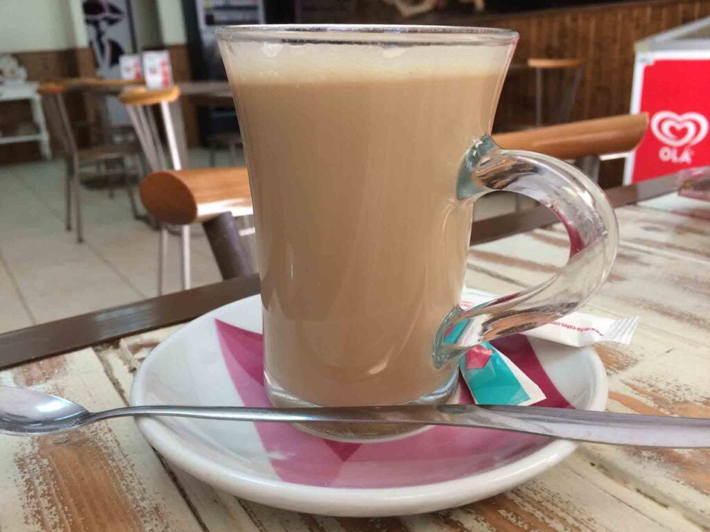 A clear glass mug filled with a creamy beige latte (called a galão in Portugal) sits on a white saucer with a pink napkin. A spoon and a small sugar packet rest beside the mug. The setting appears to be a cozy café in Portugal, with wooden chairs and tables in the background.