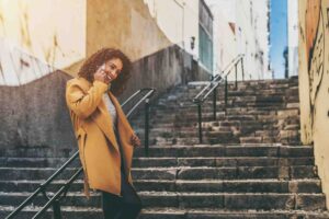 A person with curly hair, wearing a yellow coat, stands on a stone staircase and smiles while talking on the phone. Sunlight illuminates part of the scene, and the stairs are flanked by walls with graffiti.