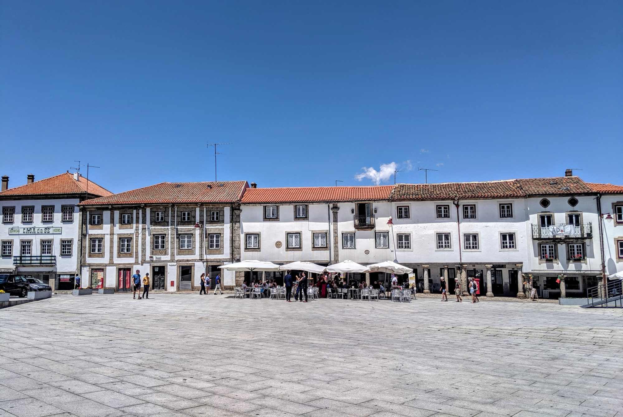 A town square in Portugal with historic-style buildings under a clear blue sky. The buildings have white facades with some stone details and red-tiled roofs. People are walking around and sitting under the white parasols of an outdoor cafe.
