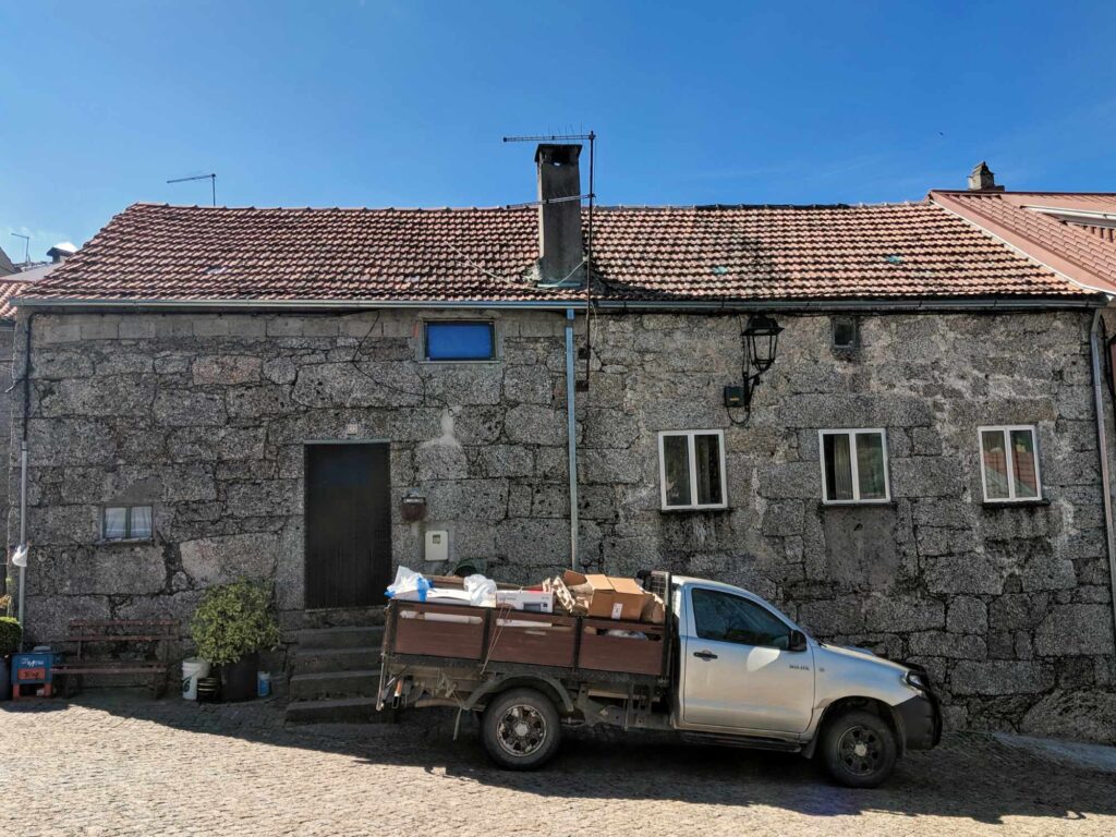 A white pickup truck loaded with boxes and miscellaneous items is parked in front of a rustic, stone-walled building with a red-tiled roof. The building has several small windows and a wooden door. Steps lead up to the door, and the sky above is clear and blue.