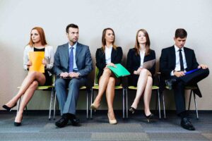 Five professionally dressed individuals sitting on chairs in a waiting area, each holding folders of different colors: orange, yellow, green, and blue. They appear to be waiting for an interview or meeting. The group includes two men and three women.