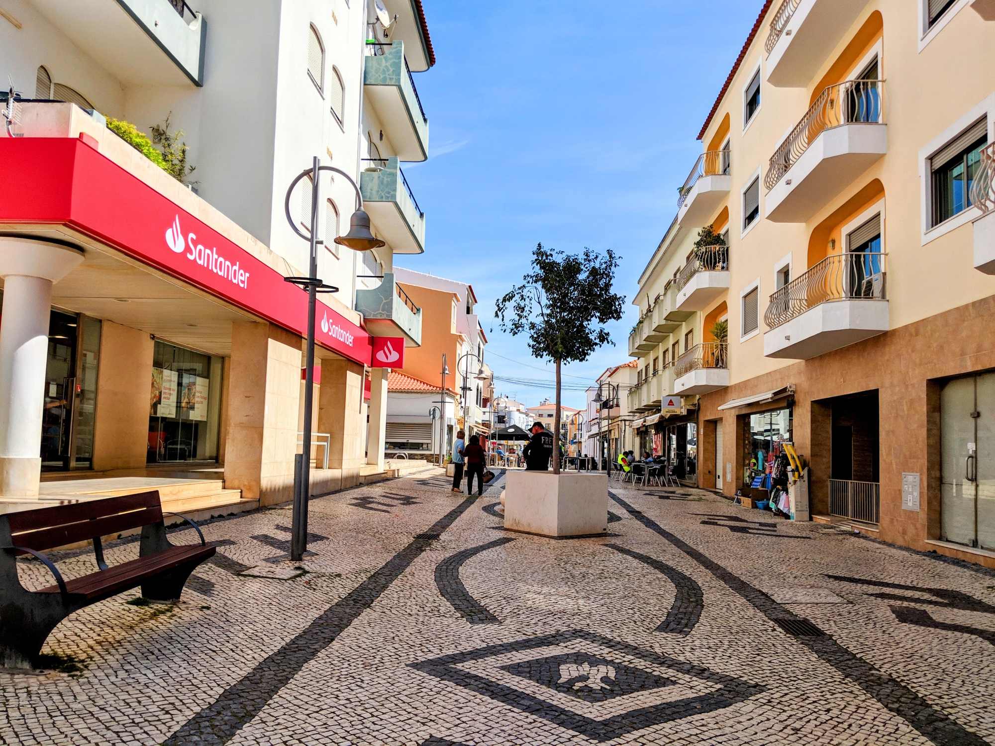 A vibrant pedestrian street in a sunny Portuguese town, lined with modern buildings, including a Santander bank on the left. The patterned cobblestone walkway features a geometric design. People are seen walking, and balconies with railings adorn the buildings.
